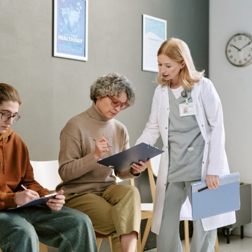 Teenager and senior woman completing medical forms while female doctor guiding paperwork in clinic waiting room. Useful for healthcare access, patient intake, family medicine marketing